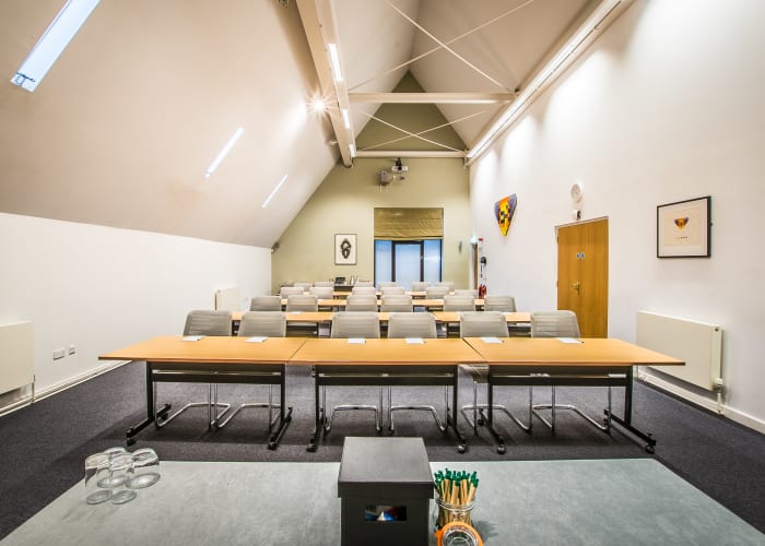 Modern tall ceiling meeting room with a slanted angled ceiling, bright space with rows of long tables with chairs and a top table at the front of the room.
