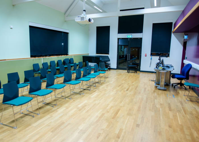 Suitable for hire, this hall has high ceilings with white metal beams, large TV screen, wooden flooring and blue chairs.
