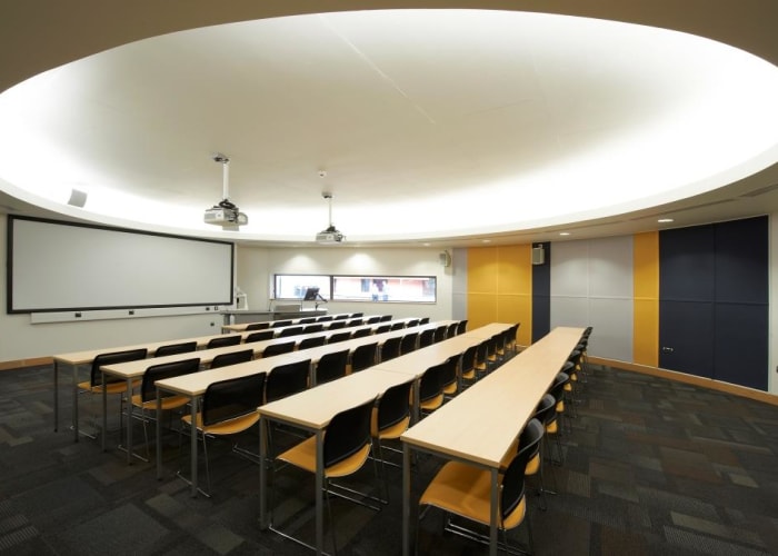 A classroom with a attractive suspended round celling with lights, wooden desks, yellow walls and chairs great for small meetings