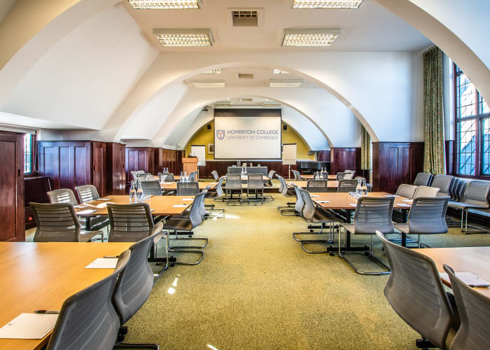Impressive meeting room in Cambridge, long room with tall ceiling, numerous square tables with chair at each table, white curved ceiling and screen at the top of the room