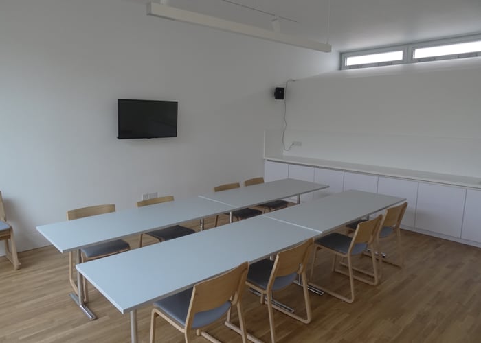 A white walled meeting room with long tables and chairs. Wall mounted TV with wooden flooring ideal for small day meetings.