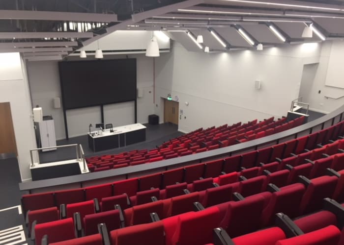 A large auditorium with rows of red uniform seats, projector and screen which is ideal for day meetings and lectures.