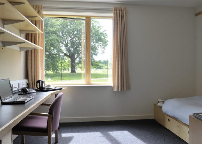 Conference bedroom with three long shelves on the wall, desk and chair. Single bed with white bedding and the room is filled with natural light and views of the extensive gardens