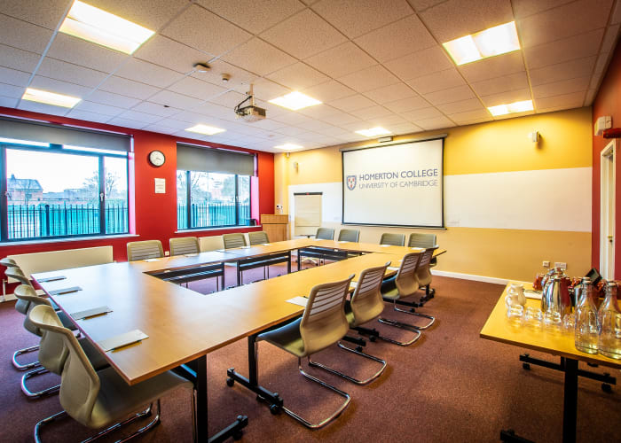 Modern event space in Cambridge, Bright room with large windows and dark red decor, set up in a hollow boardroom style and refreshments available to the side of the room and fully equipped with AV equipment.