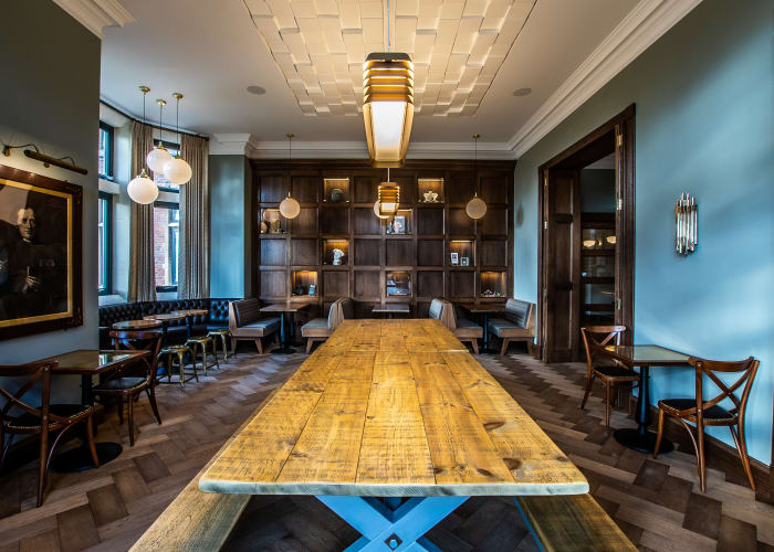 Stylish decor with a mix of blue and dark wood paneling on the walls, chrome features, modern yet also traditional area. Long wooden table taking center stage with some individual tables and chairs around the edge, ideal space to relax, socialise and have a drink.