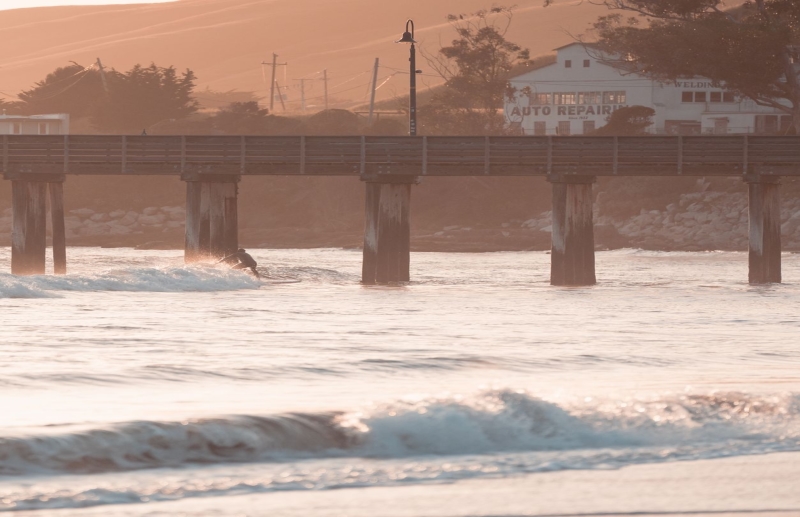 Sunset over a wooden pier with a surfer riding a wave beneath its posts; ocean waves and a coastal town harbor in the background.
