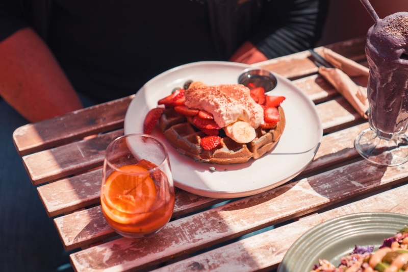 Waffle topped with strawberries and banana slices on a white plate, set on a rustic wooden table with an orange drink and a chocolate shake nearby.