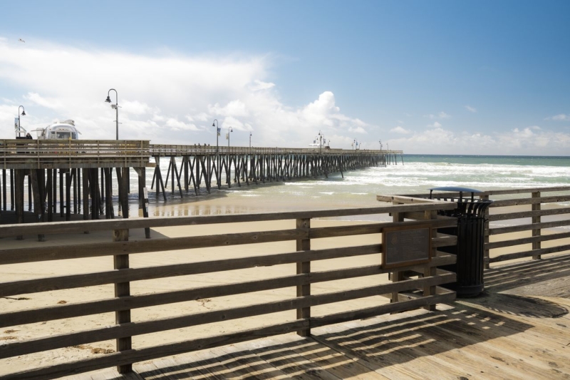 Sunny beach scene with a long wooden pier stretching into the ocean, railings along a boardwalk, a trash can, and a bright blue sky.