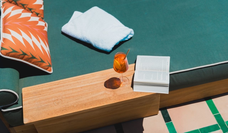 Book open next to aperal spritz on a side table next to a green pool lounger