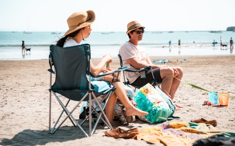 Couple relaxing on a sunny beach in folding chairs wearing wide brim hats, chatting as the ocean and other beachgoers are in the background.