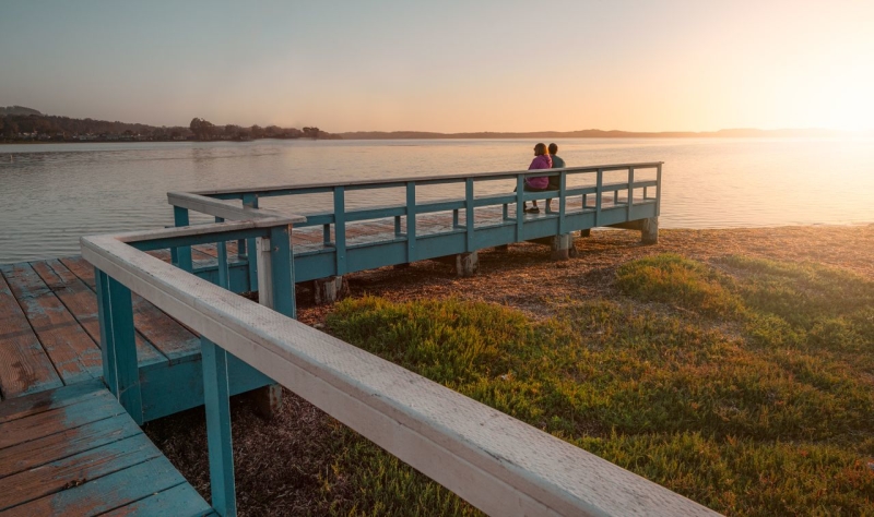 Two people sit on a blue wooden pier at sunset, watching calm water and the grassy shoreline.