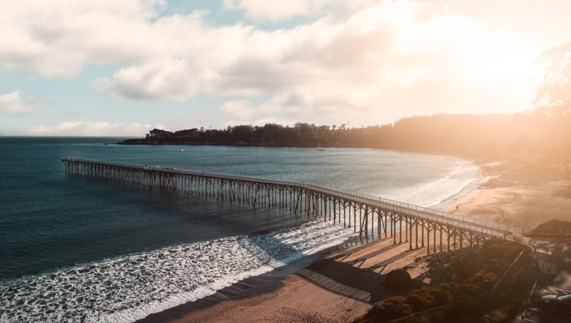 Long wooden pier stretches over blue ocean to a sandy beach at sunset, with warm sunlight, gentle waves and a California coast.