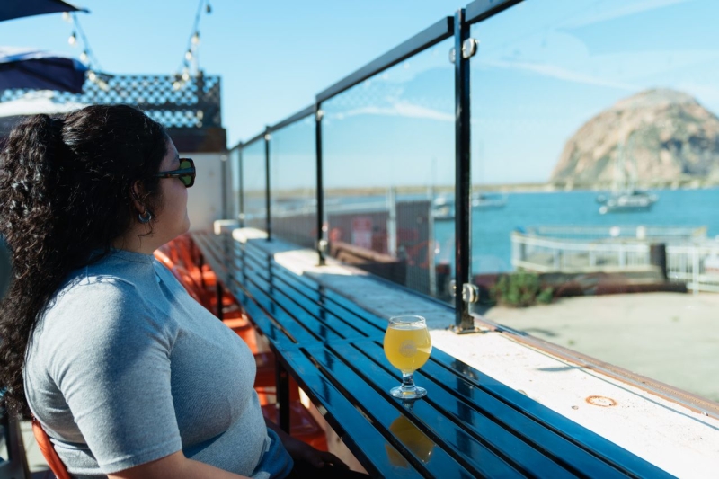 Sunlit rooftop deck with glass railing. A woman in a gray tee sits on a blue bench, a yellow drink on the table, Morro Rock visible in the distance.