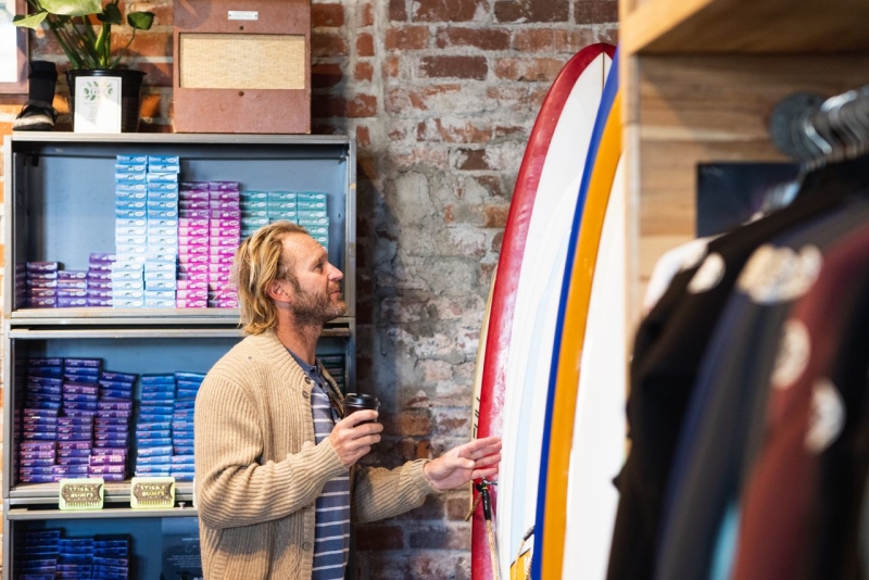 Man browsing surfboards and clothing racks with a coffee in hand