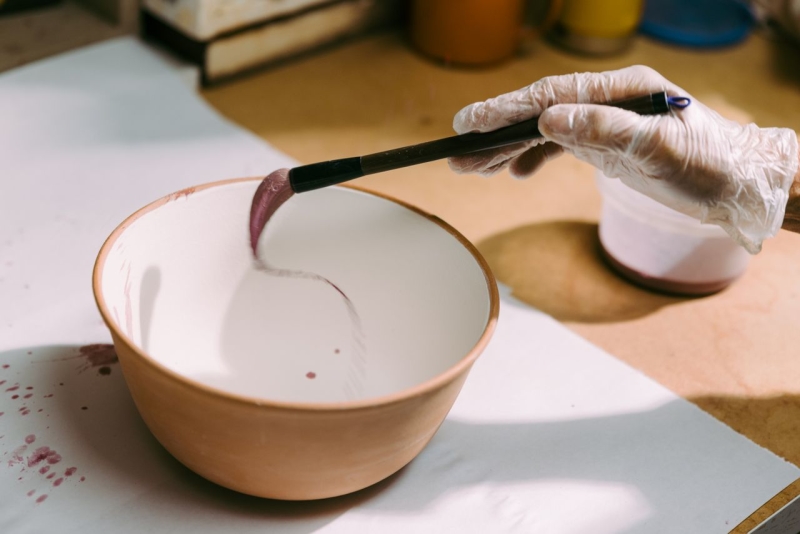 Gloved hand holding a paint brush and painting a white ceramic bowl