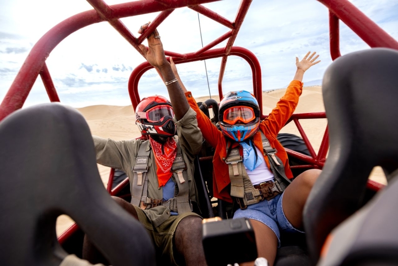 Two people with helmets and goggles in a dune buggy with hands up