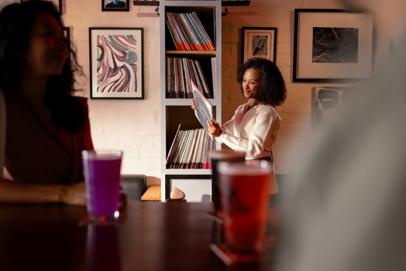 Women at bar looking at records in the background with other people in foreground with drinks