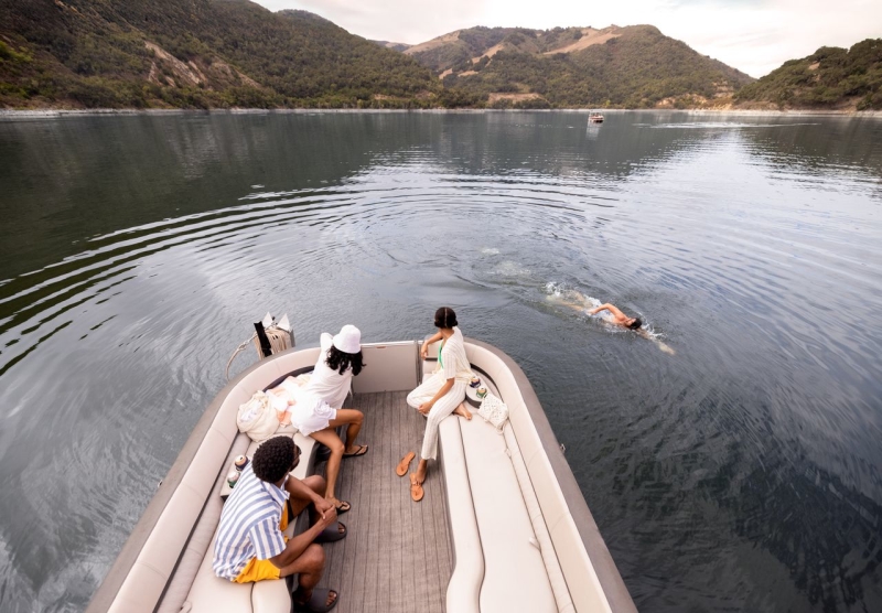 Three people in a pontoon boat watching one person swim alongside the boat in a lake