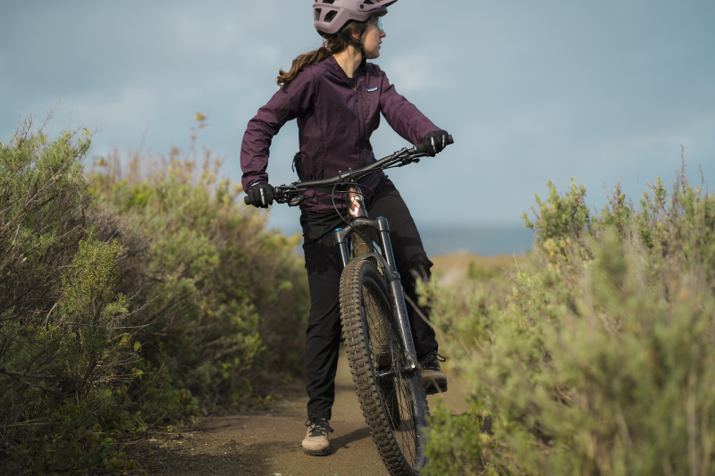 Women with purple helmet on a mountain bike stopped on trail with green bushes on either side looking out