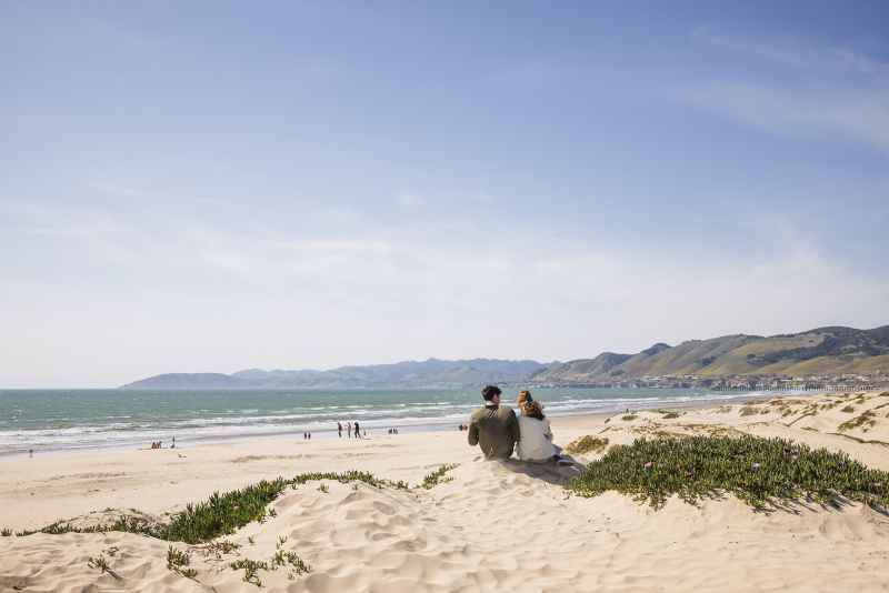 Two people sitting on the beach in Grover Beach with the coastline in the background