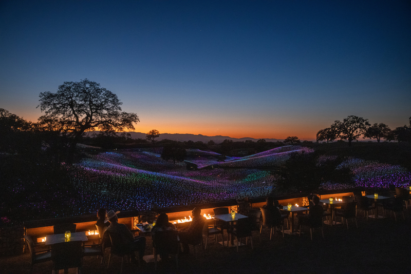 Overview of Sensorio with hills glowing and sunset with people at tables by fire in foreground
