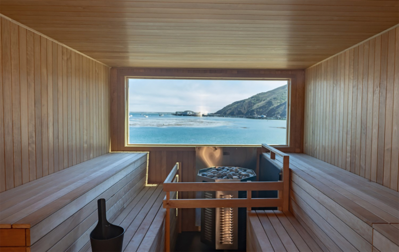 Interior view of a wood sauna with view out the window of the ocean and hill