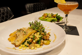 A plated trout dish with a creamy sauce and microgreens on top is next to a tall cocktail glass containing a pink drink and a smaller plate with a salad.