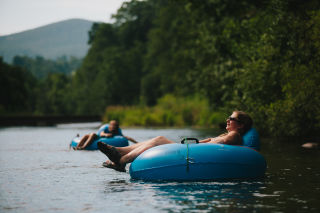 A woman relaxes in a tube with her legs hanging over the left side and her head resting on the other side. Behind her is another person in a tube with their hand in the river and a mountain in the background.