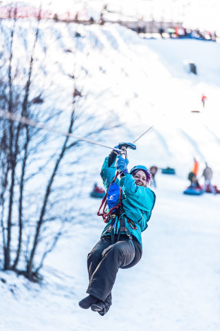 A woman ziplines above a snowy landscape, with snow tubers seen on the ground behind them.