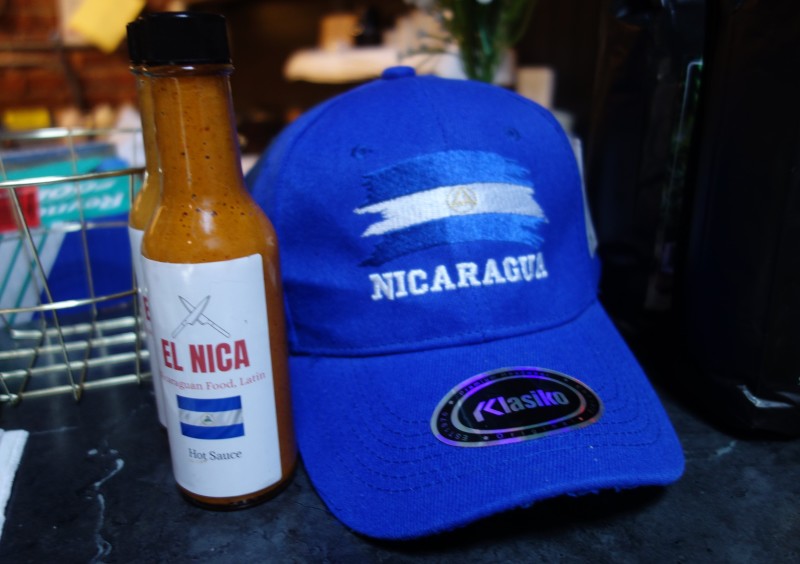                         The front counter at El Nica Spot with a bottle of hot sauce and a Nicaragua baseball hat in Hudson Valley Food Hall.
                        