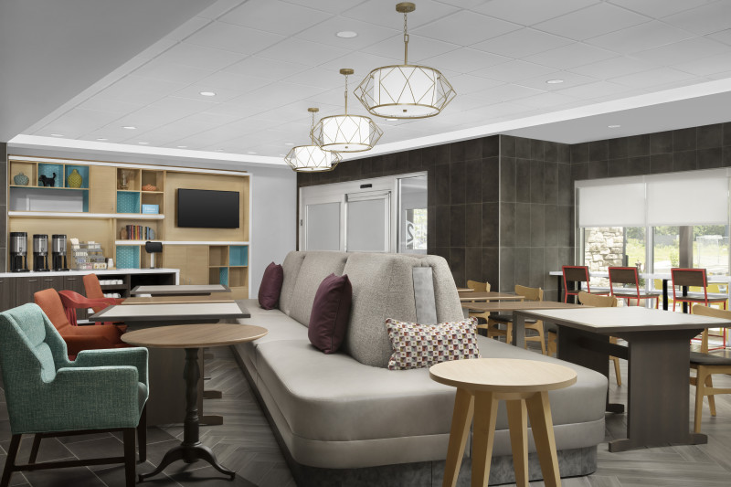                         A lounge/eating area at Home2 Suites with comfy chairs, several tables, stools, a sofa and a countertop with shelves and small kitchen appliances.
                        