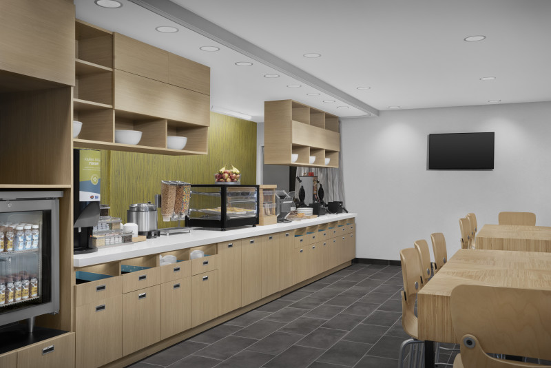                         The breakfast area at Home2 Suites with a small refrigerator with a clear door, a countertop with small kitchen appliances, along with wooden tables and chairs.
                        