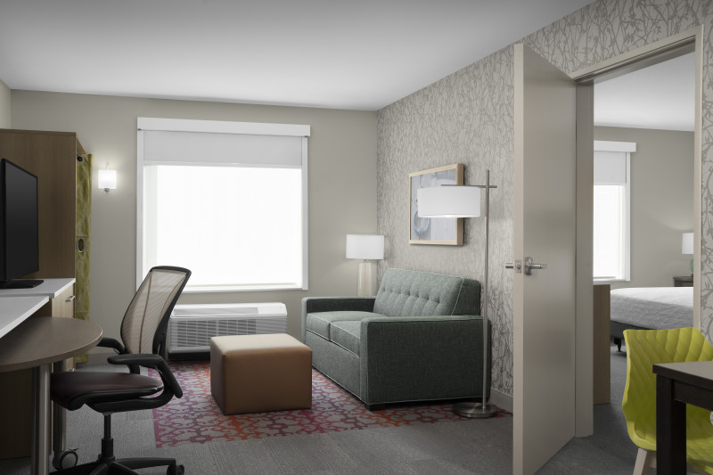                         One of the rooms at Home2 Suites, complete with a desk and chair, a table and a chair, a sofa with an ottoman, a flat-screen TV and adjoining room with a bed.
                        