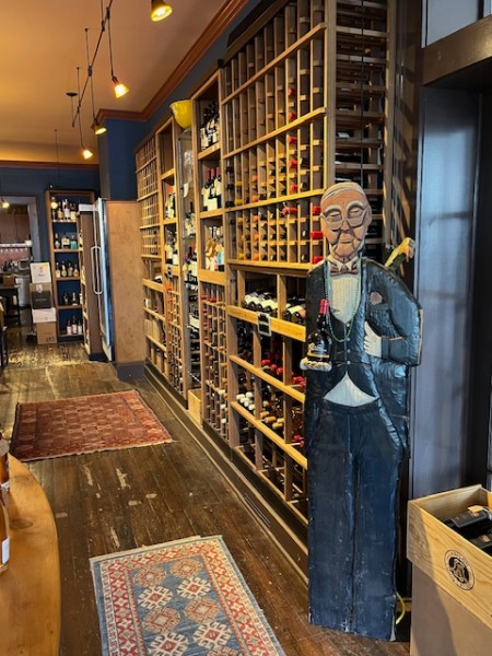                         A life-size wooden cutout of a butler in a tuxedo stands in front of the racks of bottles and holds in his hand a dark bottle with a red wax seal on top at Millerton Wine & Spirits.
                        