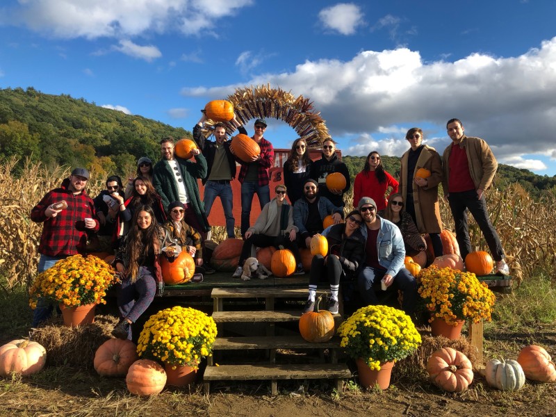                         A group of people holding pumpkins sits on a wooden porch surrounded by yellow mums at Dykeman Farm in Pawling.
                        