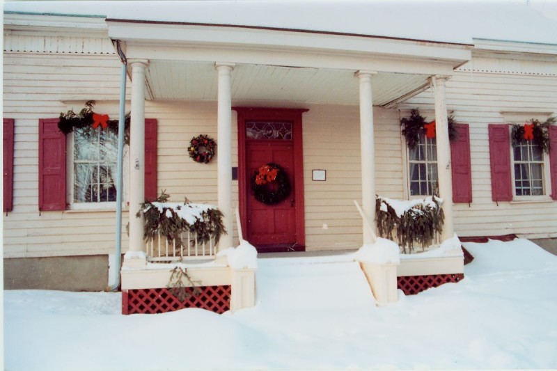                         The front of the Van Wyck Homestead, a white house with red shutters, a red front door and white columns. The home is shrouded in a layer of snow and decorated with wreaths and bows.
                        