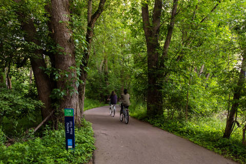 Two bikers riding on the Lansing River Trail surrounded by lush green plants