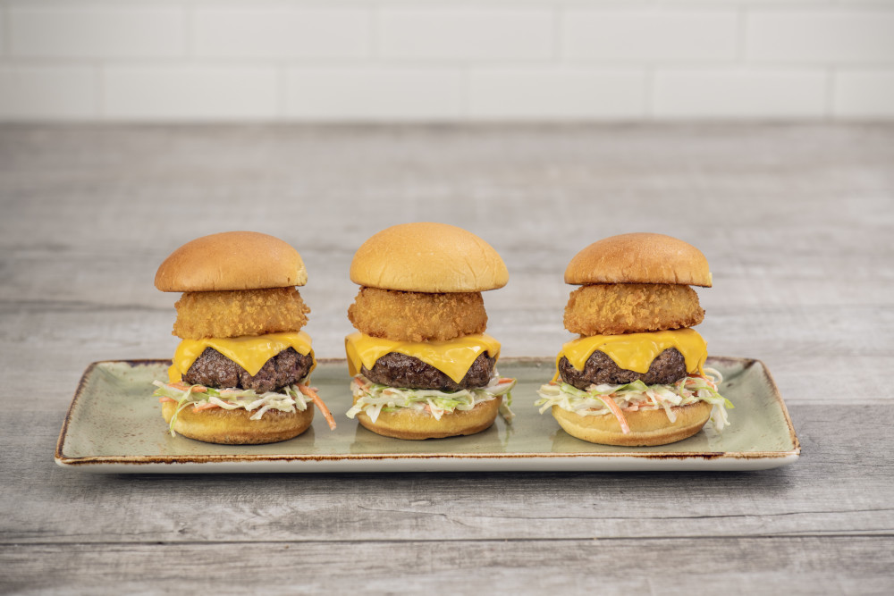 Three mini-hambúrgueres com queijo americano derretido, rodelas de cebola crocantes e salada de repolho cremosa em pão de brioche torrado.