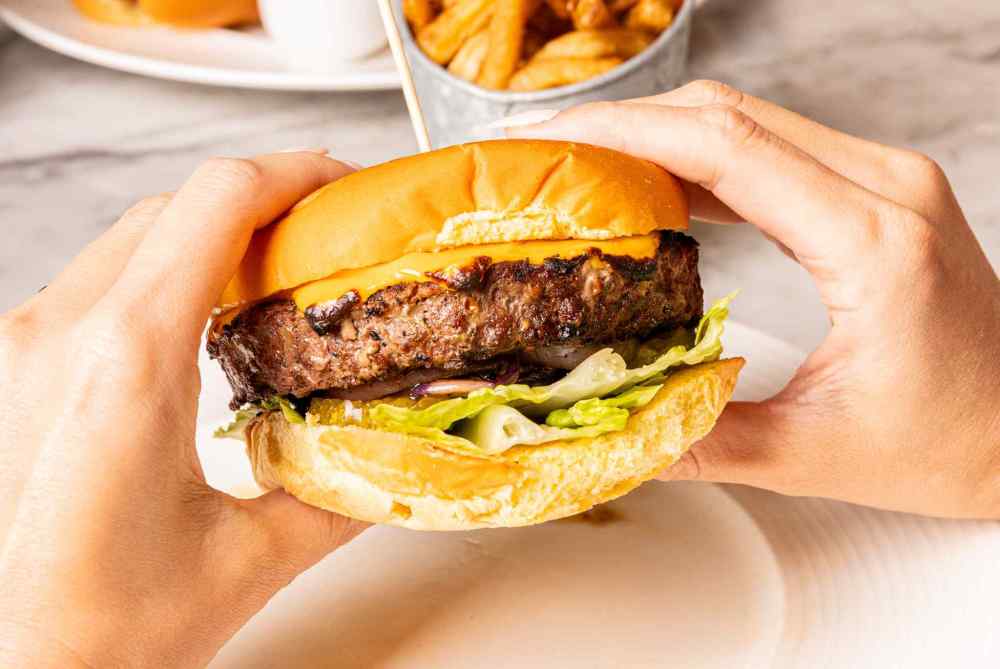 Indulge in the Wagyu Blossom Burger—Snake River Farms patty, cheddar, grilled onions, pickles, lettuce & spicy aioli. Pure handheld perfection.
