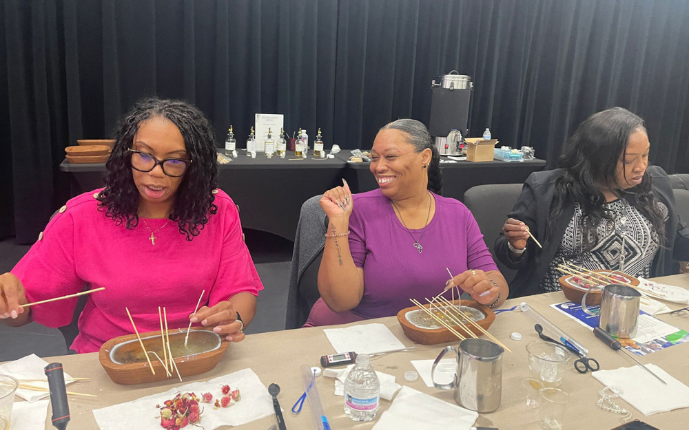 Three women at a place of aroma candle making workshop, smiling and crafting with bowls, wicks, and tools on the table.