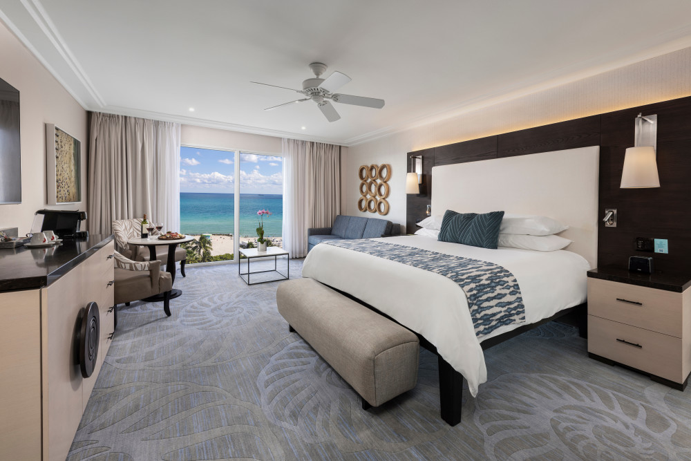 Deluxe Oceanfront King with floor-to-ceiling ocean views, a plush king bed, pull-out sofa bed, elegant coastal décor, and a relaxing seating area.