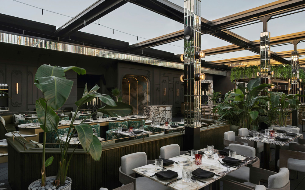 Open-air dining space features lush greenery, marble tables and elegant seating in a stylish rooftop setting