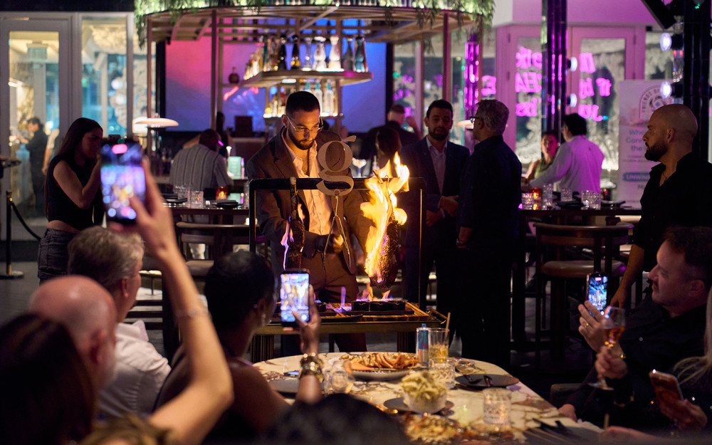A tableside dining experience at Giselle Miami featuring a dramatic flame presentation as guests enjoy dinner in the lively restaurant setting