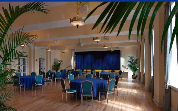 Elegant and spacious event hall featuring polished wood flooring, stylish blue-draped round tables, and gold-accented chairs.