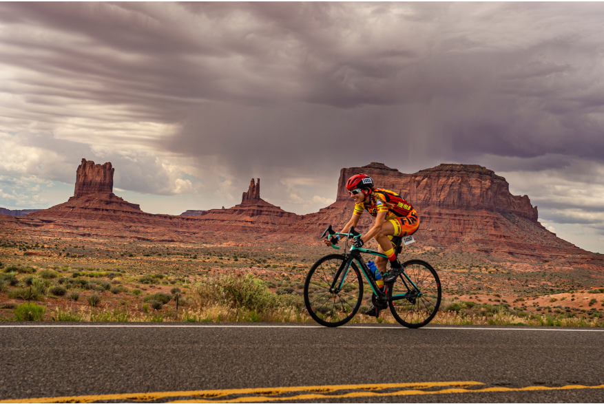 a woman rides a bike across a desert with canyons and mountains in the background