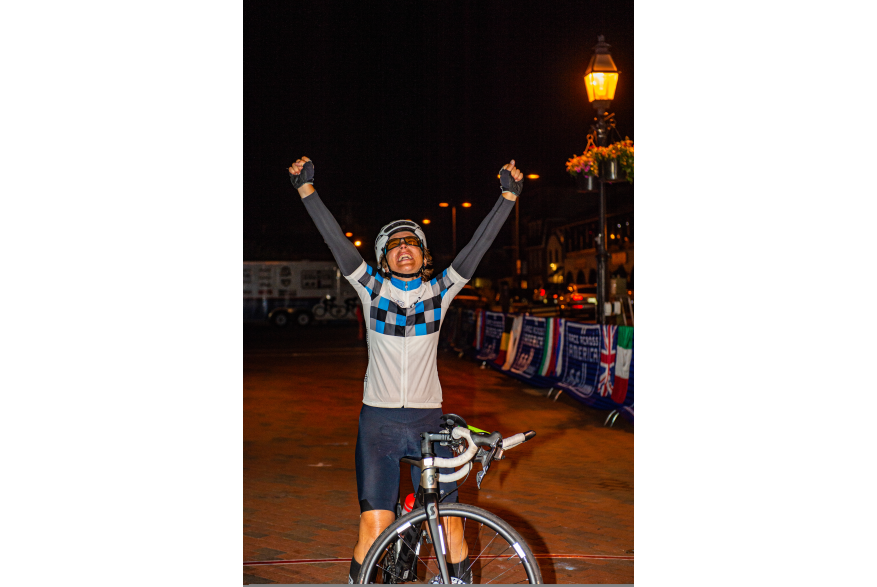 A woman with raised hands crosses the finish line on her bike at night
