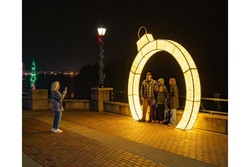 family poses for a photo under a large holiday light exhibit along a riverwalk