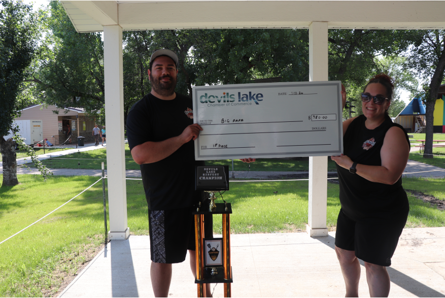 A man and a woman holding the check for first place, the trophy is orange with a plaque and is sitting on the ground.