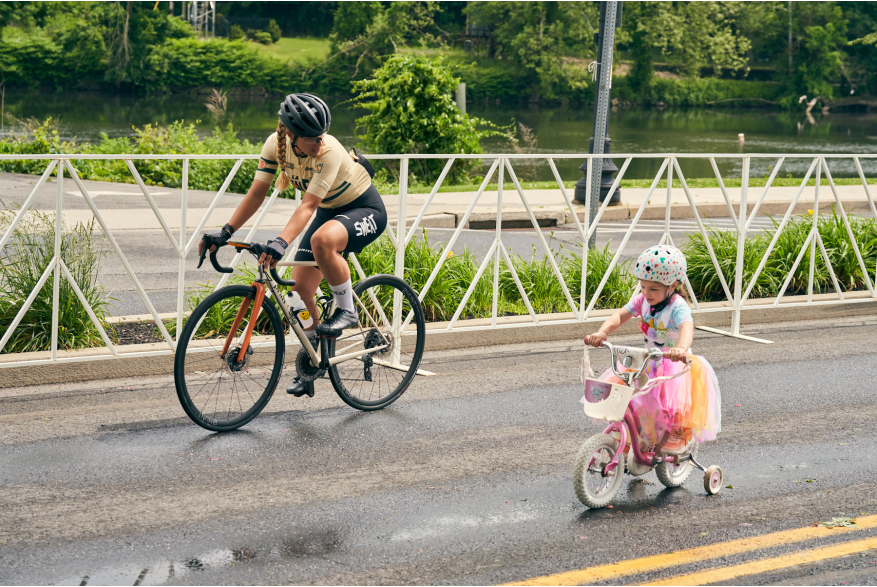 A child wearing a tutu rides her training wheels along side an adult rider at the 2022 Easton Twilight Criterium in Easton, Pa.