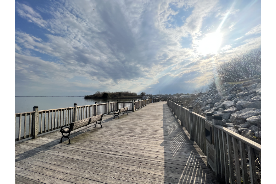 Dock walkway over water on sunny day. Benches line the way with boats in the far distance